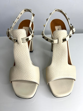 Pair of white textured high-heeled sandals with metallic straps on a light gray background