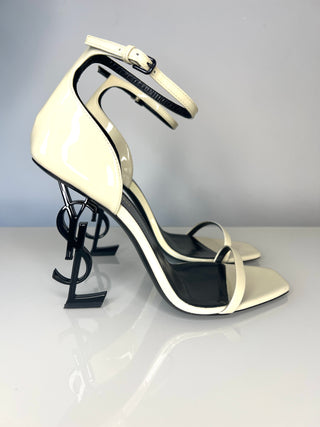 A pair of Saint Laurent Opyum patent leather high-heeled sandals in cream and black, featuring a unique sculptural black metal YSL heel and an ankle strap.