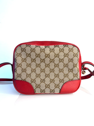 A Gucci crossbody camera bag featuring the classic beige and brown GG monogram canvas on the front, with vibrant red leather trim and a matching red shoulder strap