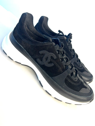 A pair of black Chanel sneakers with a black and white sole, featuring the iconic interlocking CC logo on the side.