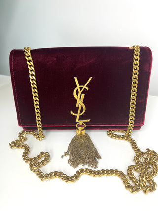 A Saint Laurent shoulder bag in deep burgundy velvet with a gold-tone YSL logo clasp and a matching gold chain strap with a tassel.