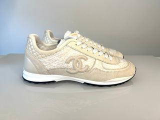A pair of Chanel low-top sneakers in a light beige and cream colorway, featuring textured leather panels, quilted details on the heel, and the signature interlocking CC logo on the side.