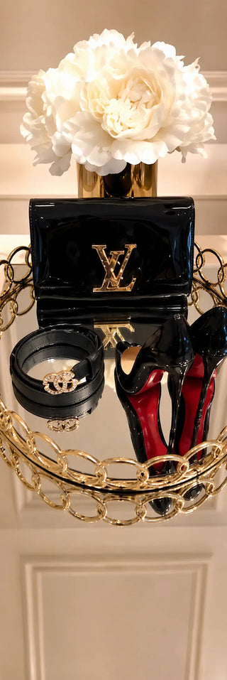 A glossy black Louis Vuitton wallet with a gold LV logo, styled on a mirrored tray with a black Gucci belt, Christian Louboutin black high heels, and white flowers against a beige wall.
